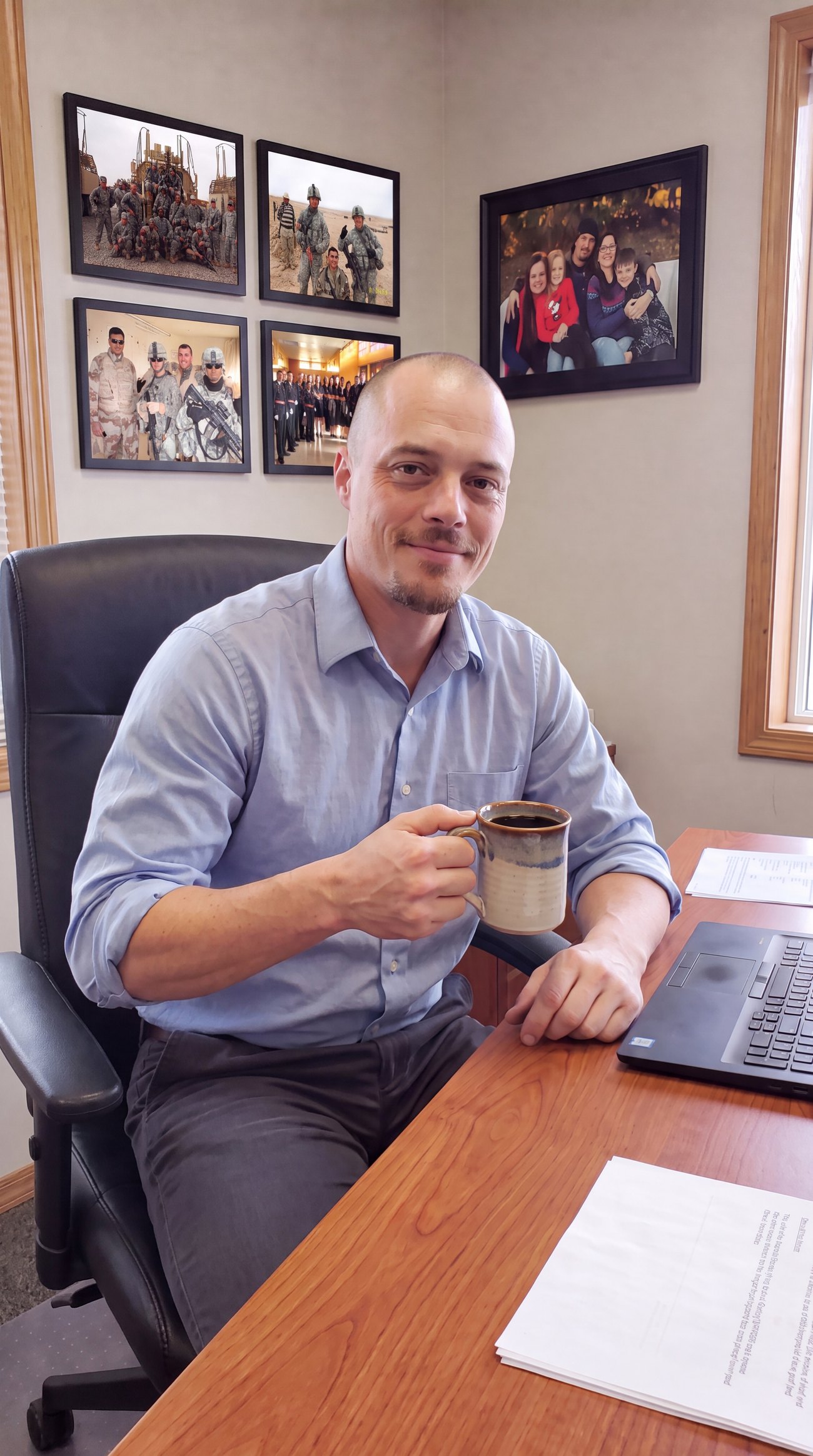 Bruce McMahan, Founder and disabled Veteran, at his desk with a cup of coffee, his military service photos and family photo framed on the wall behind him